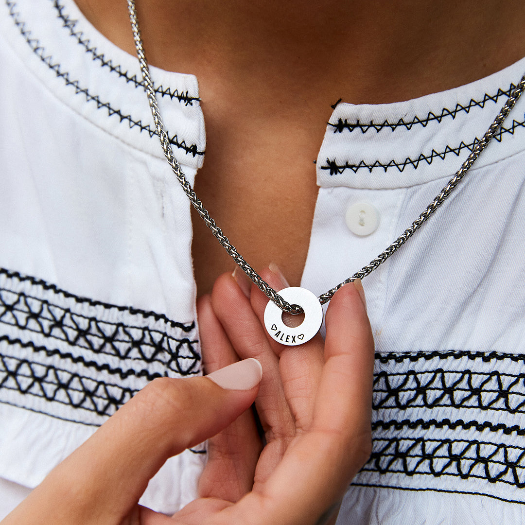 Mano sosteniendo la Anilla® grabada del collar Native sobre una blusa blanca